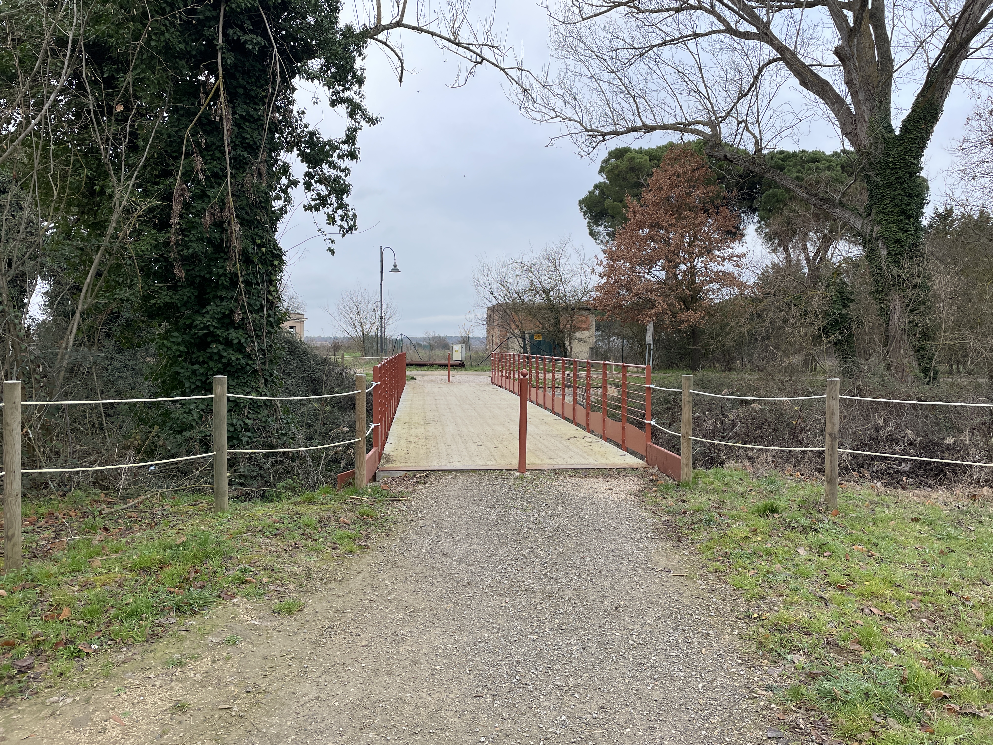 Bicycle-pedestrian bridge with red railings over water course. Gravel path edged with white ropes